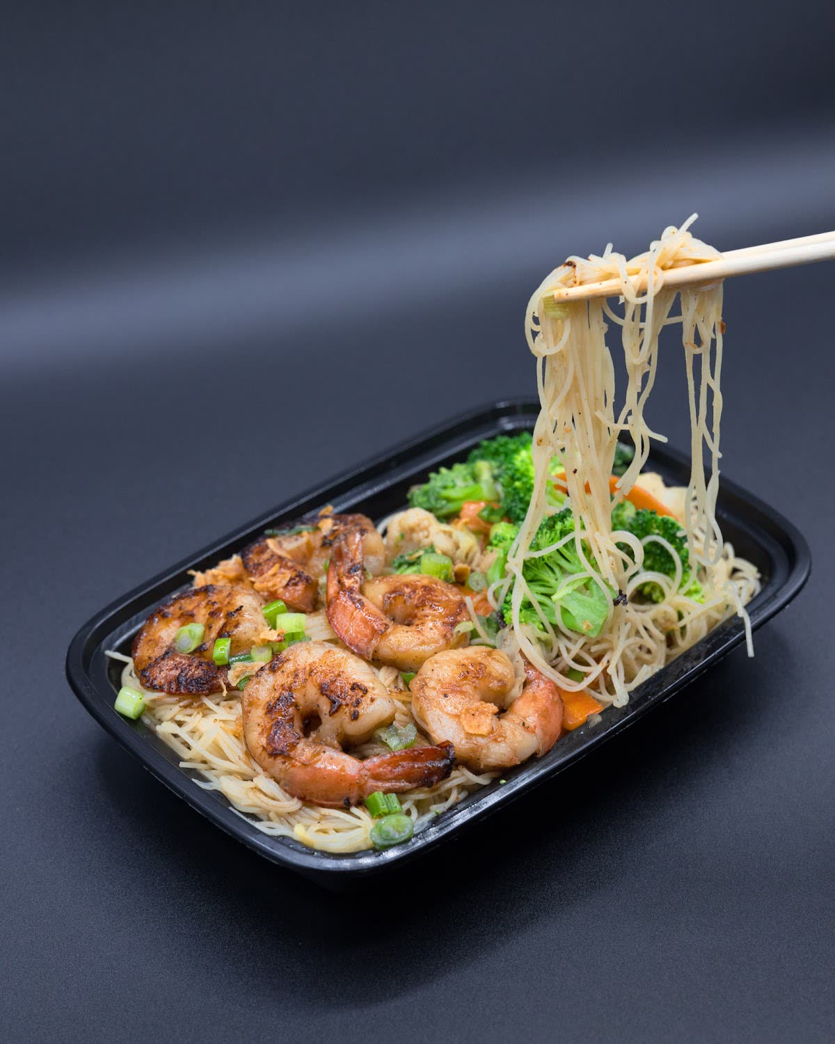 Chopsticks lifting noodles from a tray of shrimp and vegetables