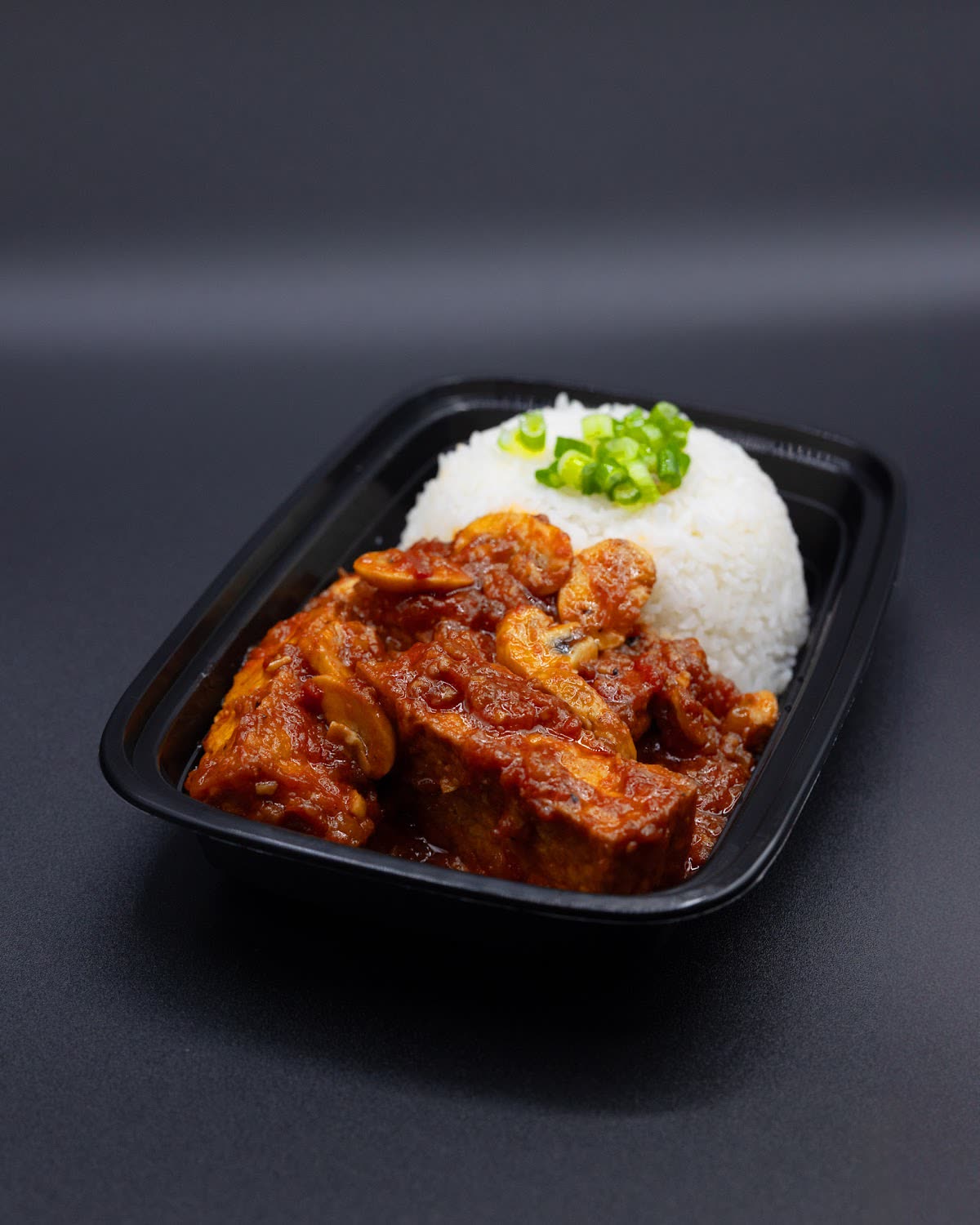 Black takeaway tray with rice and sauced meat against a dark background
